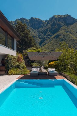 Dağlara bakan büyük havuzlu modern iki katlı bir ev. Tatilin tadını çıkarmak için iki güneşli yatak ve geniş bir şemsiye. İçeride kimse yok.