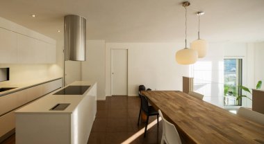 Yuvasının üstünde kapüşonu olan modern bir mutfak. Yakınında üzerinde iki güzel lamba olan büyük bir ahşap masa var. Pencereden içeri bir ışık huzmesi giriyor..