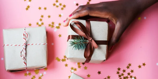 African womans hand taking a craft textured gift box with ribbon bow surrounded by stars and snowflakes on a coral background, from above — 图库照片