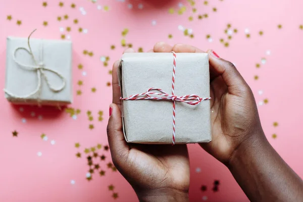 Mãos de mulher africana segurando caixa presente com um arco no fundo rosa pastel com estrelas e flocos de neve confetti. Foco seletivo — Fotografia de Stock