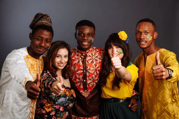 Mírumilovná koncepce přátelství mezi zeměmi: tři Afričané a dvě latinskoamerické dámy v národních šatech ukazují palec nahoru objetí spolu s vlajkami tělesného umění Kongo, Nigérie, Brazílie, Ekvádor — Stock fotografie