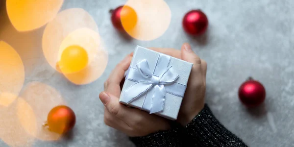 Las manos de las mujeres están sosteniendo una caja de regalo con textura artesanal con lazo de cinta por bolas de Navidad vinosas desenfocadas sobre un fondo gris con destello de guirnalda, desde arriba —  Fotos de Stock