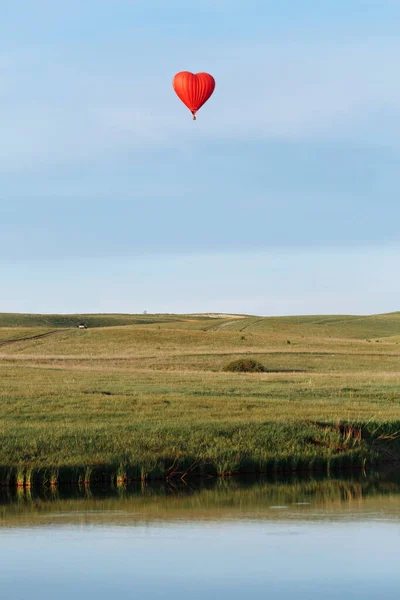 Hot air balloon in the shape of a heart is landing behind the lake, mirroring in it — Stock Photo, Image