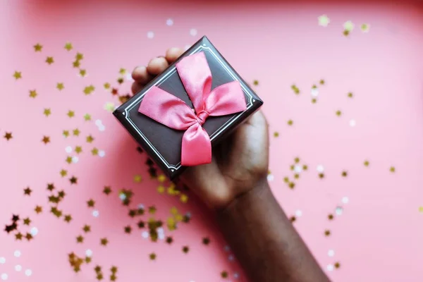 Рука африканки тримає коробку з луком на рожевому фоні з зірками і сніжинками конфетті. Вибіркове зосередження — стокове фото