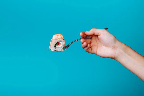 Mão segurando rolo de sushi kunsan fresco com um garfo, isolado em fundo azul. Como comer sushi sem pauzinhos — Fotografia de Stock