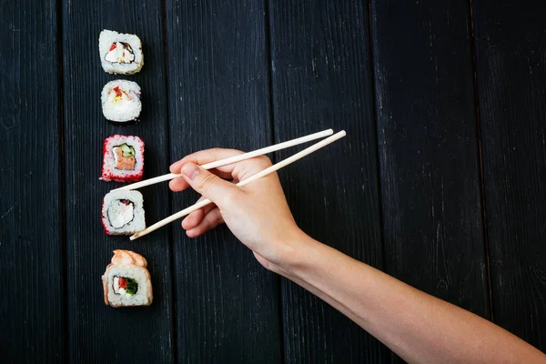 Mão feminina leva pauzinhos rolos de sushi com pauzinhos chineses. Deitado numa tábua de madeira preta. Vista de cima — Fotografia de Stock