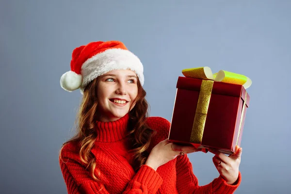 Piękna Europejka w kapeluszu Mikołaja trzyma prezenty, patrzy w kamerę i uśmiecha się, odizolowana na szarym tle — Zdjęcie stockowe