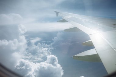 Beyaz kabarık bulutların ve mavi gökyüzünün üzerinde kanatları olan uçak penceresinden bak.