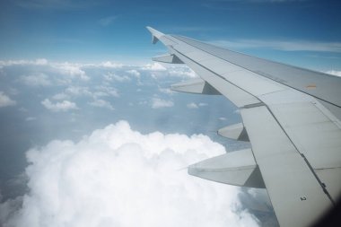 Beyaz kabarık bulutların ve mavi gökyüzünün üzerinde kanatları olan uçak penceresinden bak.