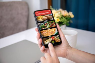 Kadın eli, evdeki odanın arka planına karşı ekranda yemek dağıtım uygulaması olan telefonu tutuyor.