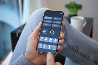 Erkek eller, odasındaki ekranda akıllı ev uygulaması olan telefonu tutuyor. 