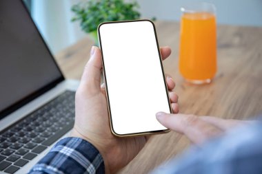 Masanın arka planında, odada dizüstü bilgisayarı olan yalnız ekranlı telefonu tutan erkek eller.