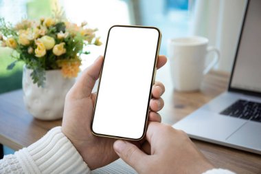 Masanın arka planında, odada dizüstü bilgisayarı olan yalnız ekranlı telefonu tutan erkek eller.