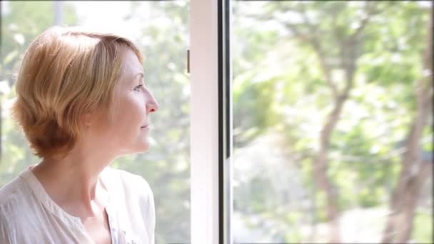 Uma mulher madura atraente fica na janela da imagem admirando a natureza — Vídeo de Stock
