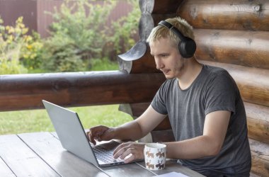 Kulaklıklı bir adam dizüstü bilgisayarında bir şey yazıyor, bir kır evinin terasında oturuyor.