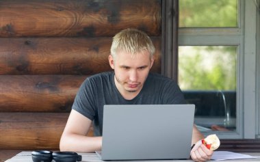 Adam bir kır evinin terasında oturup dizüstü bilgisayarla çalışmaya odaklanmış.