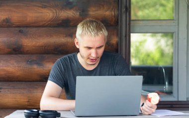 Adam bir kır evinin terasında oturup dizüstü bilgisayarla çalışmaya odaklanmış.