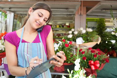 florist woman using the phone with a clipbard in her store