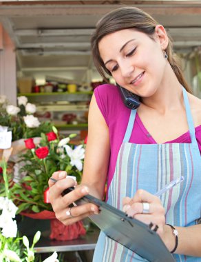 florist woman using the phone with a clipbard in her store