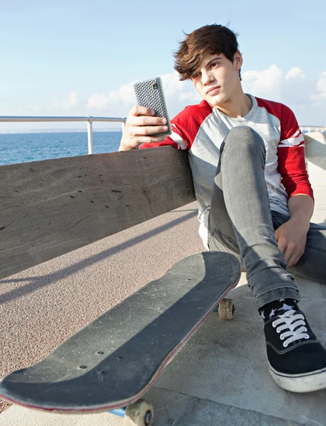 Menino com um skate segurando um smartphone em um banco — Fotografia de Stock