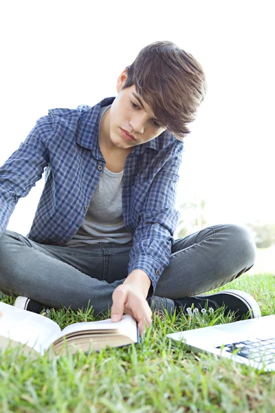 Menino em um parque lendo um livro e usando um laptop — Fotografia de Stock