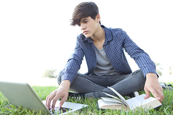 Junge im Gras liest ein Buch und benutzt einen Laptop — Stockfoto