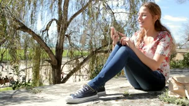女性が公園で彼女のスマート フォンのイヤホンで音楽を聴く — ストック動画