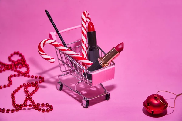 Shopping cart with red lipstick, candy cane, mascara. On a pink background. With a red Christmas ball and beads. Concept of Christmas shopping for beauty products.