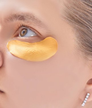 Göz altı kolajen altın pedli bir kadın. Cilt bakımı yapan kız göz torbalarını küçültmek için göz altındaki kumaş maske parçalarına dokunuyor. Cilt bakımı konsepti, yaşlanmayı önleyen nemlendirici göz maskesi, altın hidrojel bantlar..