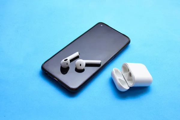 Russia, Udachnyy, April 20, 2021. White wireless bluetooth earphones or headphones, plastic case or box for storage and charging and smartphone, on color background close up
