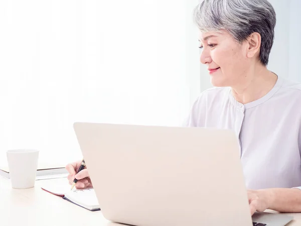 Modern elderly grey-haired 60s woman take online course or training on computer at home.