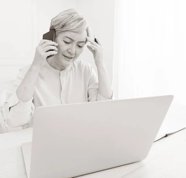 Asian senior woman at phone works on her laptop at home.