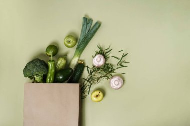 Yeşil sebzeler ve meyveler açık yeşil arka planda bir kesekağıdından dökülüyor..