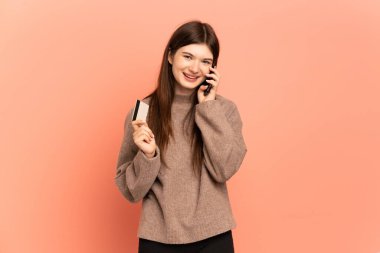 Pembe arka planda izole edilmiş genç Ukraynalı bir kız cep telefonuyla sohbet ediyor ve elinde bir kredi kartı tutuyor.