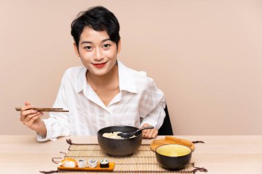 Genç Asyalı kız bir kase erişte ve suşiyle masada.