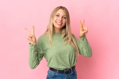 Pembe arka planda izole edilmiş genç sarışın kadın iki eliyle zafer işareti gösteriyor.