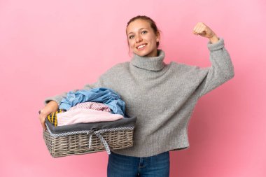 Pembe arka planda izole edilmiş çamaşır sepetini tutan genç bir kadın güçlü bir jest yapıyor.