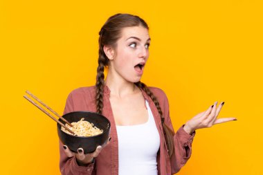 Elinde yemek çubuklarıyla bir kase erişte tutarken yüzünde sürpriz bir ifade olan izole edilmiş sarı arka planda genç bir kadın.