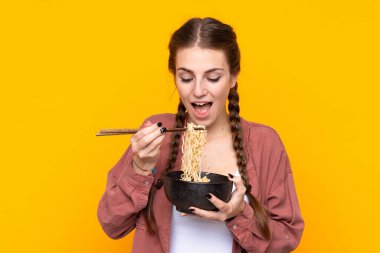 İzole edilmiş sarı arka planda genç bir kadın elinde yemek çubuklarıyla bir kase erişte tutuyor.