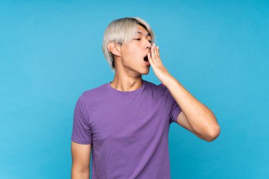 Asyalı genç adam izole edilmiş mavi arka planda esniyor ve ağzını eliyle açık tutuyor.