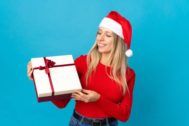 Noel şapkalı kadın elinde hediyesiyle mavi arka planda mutlu bir ifadeyle izole edilmiş.