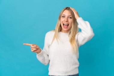 Mavi arka planda izole edilmiş genç sarışın kadın şaşırmış ve işaret ediyor.