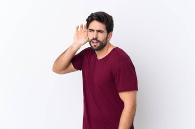 Sakallı, sakallı, beyaz arka planda genç yakışıklı bir adam. Kulağına elini sürerek bir şeyler dinliyor.