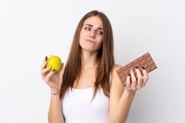 Bir elinde çikolata tableti, diğer elinde elma tutarken, beyaz arka planda kuşkuları olan genç bir kadın.