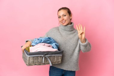 Pembe arka planda izole edilmiş çamaşır sepetini tutan genç bir kadın mutlu ve parmaklarıyla dört tane sayıyor.
