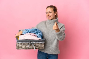 Pembe arka planda izole edilmiş çamaşır sepetini tutan genç bir kadın para hareketi yapıyor.