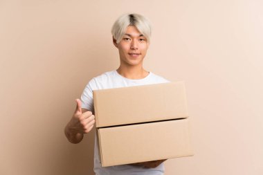 Asyalı genç bir adam izole edilmiş bir arkaplanda bir kutu tutuyor ve başparmağı yukarıda başka bir yere götürüyor.