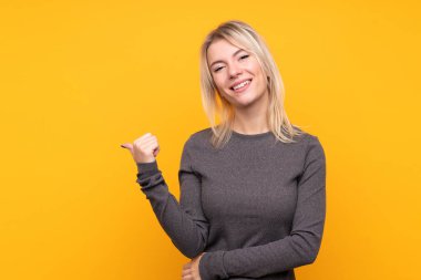 Sarışın, izole edilmiş sarı arka planda genç bir kadın bir ürün sunmak için kenarı işaret ediyor.