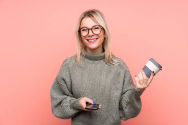 Soyutlanmış pembe arka planı olan genç, sarışın bir Rus kadın elinde kahve ve bir cep telefonu tutuyor.