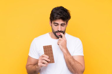 Sakallı yakışıklı genç adam izole edilmiş sarı arka planda çikolata tableti alıyor ve kuşkuları var.